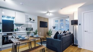 Eine moderne Küche mit weißen Schränken, ein Essbereich mit Holztisch und -stühlen, ein dunkelgraues Sofa und eine Stehlampe.