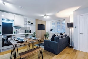 A modern kitchen with white cabinetry, a dining area with a wooden table and chairs, a dark grey sofa, and a floor lamp.
