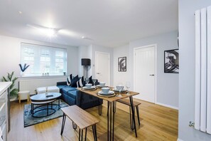 A modern living room with a sofa, a dining table set for four, and a large window with blinds.