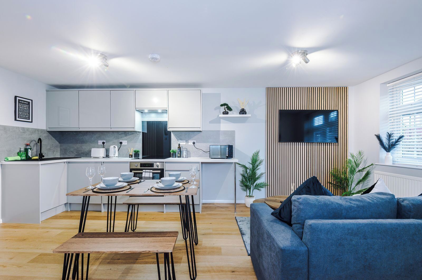 A modern kitchen with white cabinets, a dining area with a wooden table and chairs, and a blue sofa in a living room.