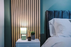 A neatly made bed with white linens, a bedside lamp, a small potted plant, and a nightstand with a lamp and a bottle.