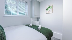 Ein Schlafzimmer mit Bett, einem Fenster mit Jalousien, einem Nachttisch, einem Spiegel und einem gerahmten Bild an der Wand.