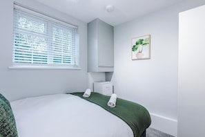 A bedroom with a bed, a window with blinds, a bedside table, a mirror, and a framed picture on the wall.