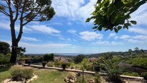 A landscaped garden with a large tree, rock planters, and a view of a coastal town. - Le Mas des iris, large swimming pool, panoramic sea view, Cap d'Antibes beaches 5 minutes away (Antibes)