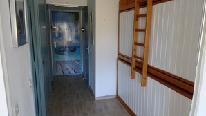Een gang met een houten vloer, een deur met een blauw kozijn, en een houten ladder die tegen een witte muur staat.