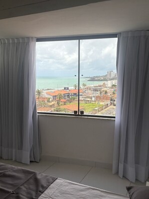 A bedroom with a large window, white curtains, a bed, and a view of a coastal cityscape. - MoviHosts Ayambra (Natal)