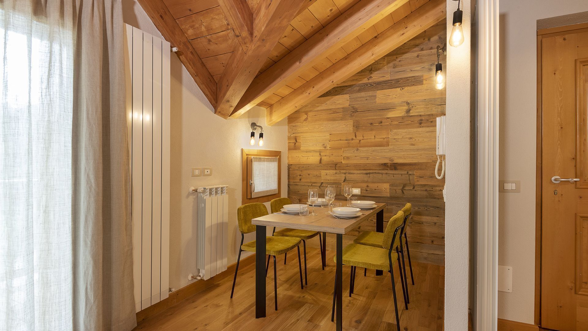 Comedor con mesa de madera dispuesta para cuatro personas, dos sillas, un radiador, una lámpara fijada a la pared y una puerta.