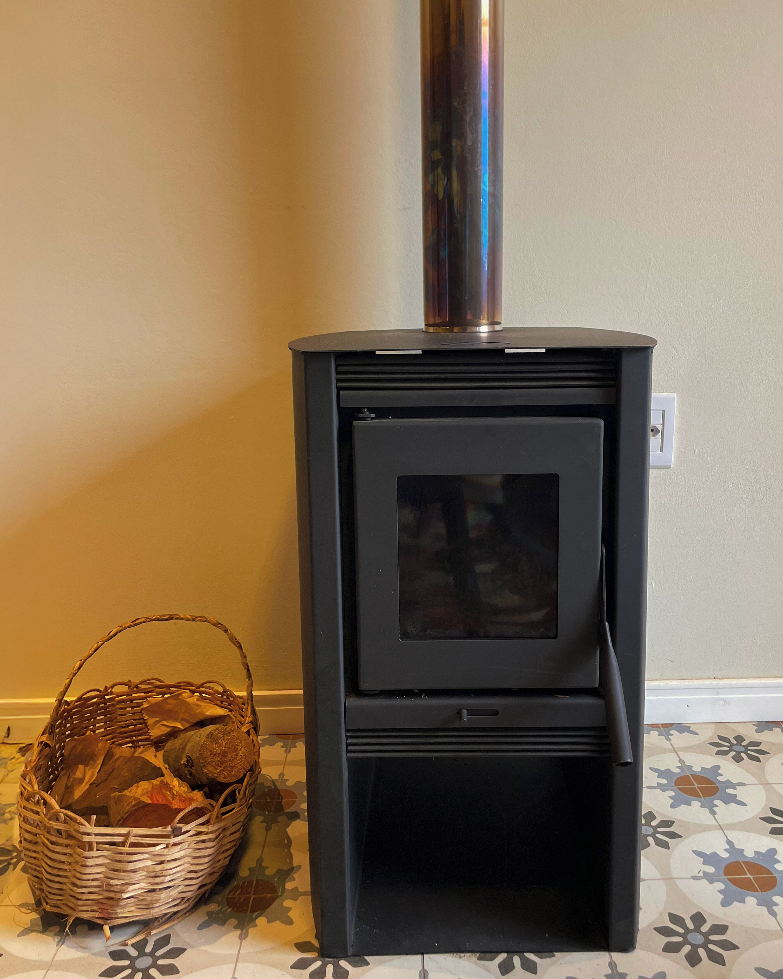 A black wood-burning stove with a glass door, a basket of firewood, and a patterned floor.