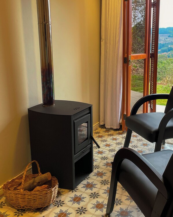 Habitación con una estufa de leña negra, una cesta de mimbre, una silla y vistas a través de un ventanal.