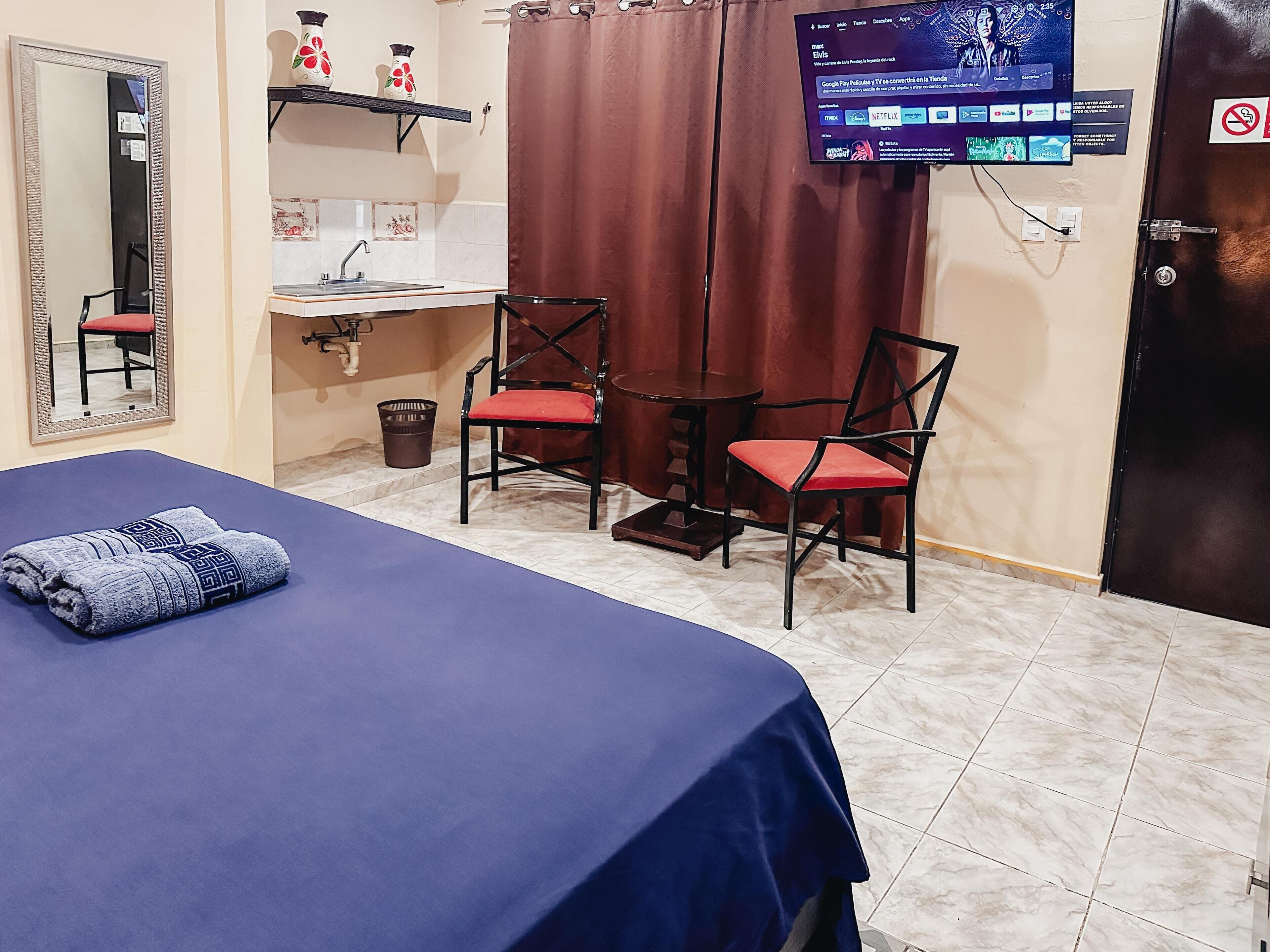 A hotel room with a bed, two chairs, a small table, a sink, and a TV mounted on the wall.