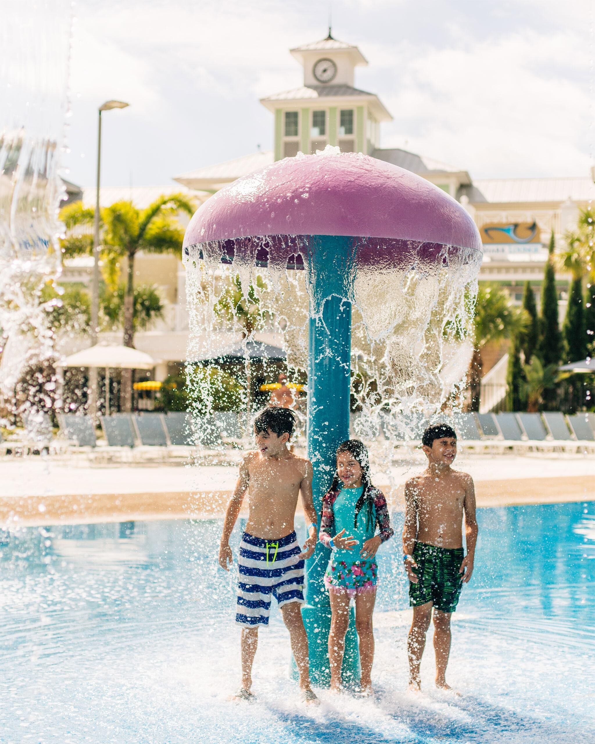 Una fuente de agua con una parte superior morada, rodeada de una zona de piscina con sillones reclinables y una torre del reloj al fondo.