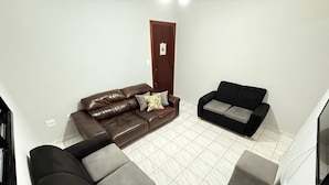 A living room with a brown leather sofa, a black sofa, and a grey ottoman. There is a wooden door with a small decoration above it. - Pé na Areia Canto do Forte VI (Praia Grande)