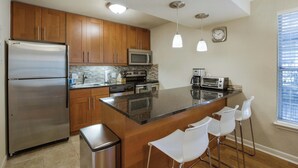 Cocina moderna con armarios de madera, electrodomésticos de acero inoxidable, encimera de granito y una zona de bar con taburetes blancos.