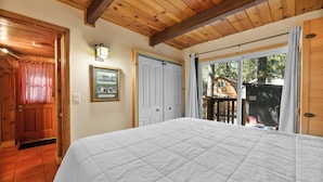 Un dormitorio con una cama grande, vigas de madera en el techo, una puerta con cortina de cuadros rojos y una puerta corredera de vidrio que da a un área exterior.