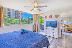Un dormitorio con una cama grande, ventilador de techo, televisor y balcón con vistas a zonas verdes y edificios.