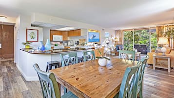 A modern kitchen with a central island, dining area with a wooden table and chairs, and a living room with a sofa and a large window overlooking greenery.
