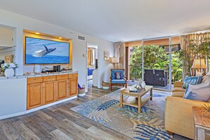 A spacious living area with a large flat-screen TV, wooden cabinets, a sofa, and a dining table. - West Maui Ocean View Corner 2BR with Pool + AC (Lahaina)
