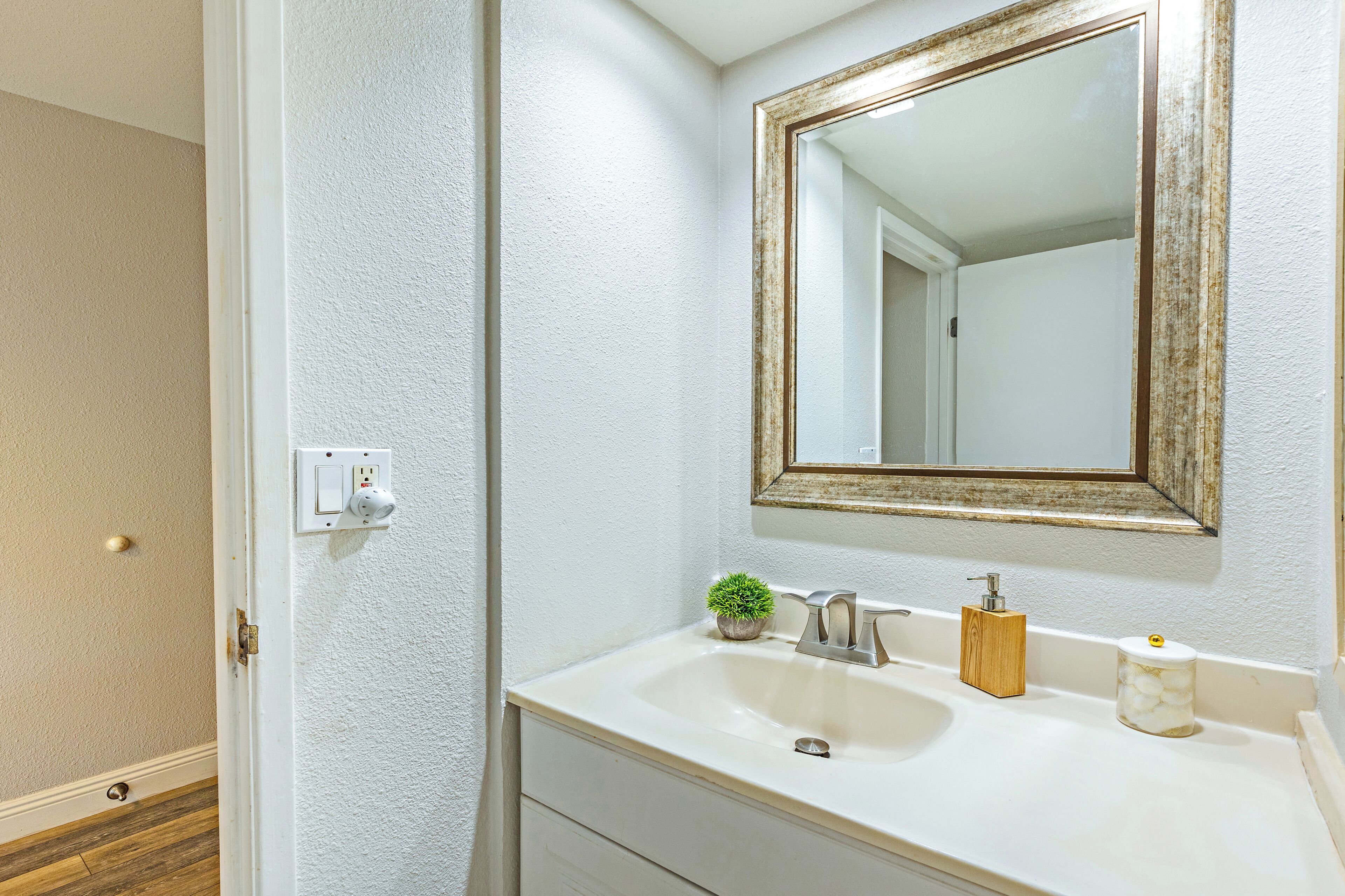 Un baño con un lavamanos blanco, un espejo con marco de madera, una pequeña planta en maceta, un dispensador de jabón y un recipiente con algodón.