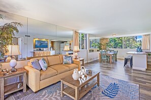 Amplia sala de estar con un sofá, una mesa de centro y una mesa de comedor. Se ve una cocina al fondo, y hay un ventanal con cortinas.