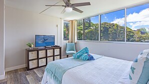 Ein Schlafzimmer mit Bett, Fernseher, Deckenventilator, Fenster mit Blick auf Grünflächen und einer Kommode mit Schubladen.