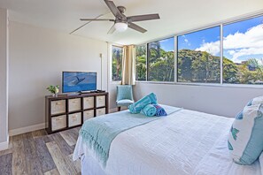 Un dormitorio con cama, televisión, ventilador de techo, ventana con vistas a la vegetación y un mueble de almacenamiento con cajones.