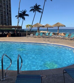 A pool area with lounge chairs and umbrellas, a clear view of the ocean, and a tall building in the background. - West Maui Ocean View Corner 2BR with Pool + AC (Lahaina)