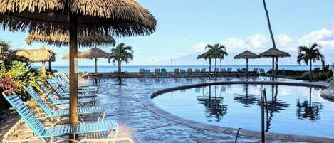 A pool area with lounge chairs and thatched umbrellas, surrounded by palm trees and a view of the ocean.