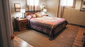 Un dormitorio con cama, mesitas de noche, un tapete estampado y dos ventanas con cortinas.