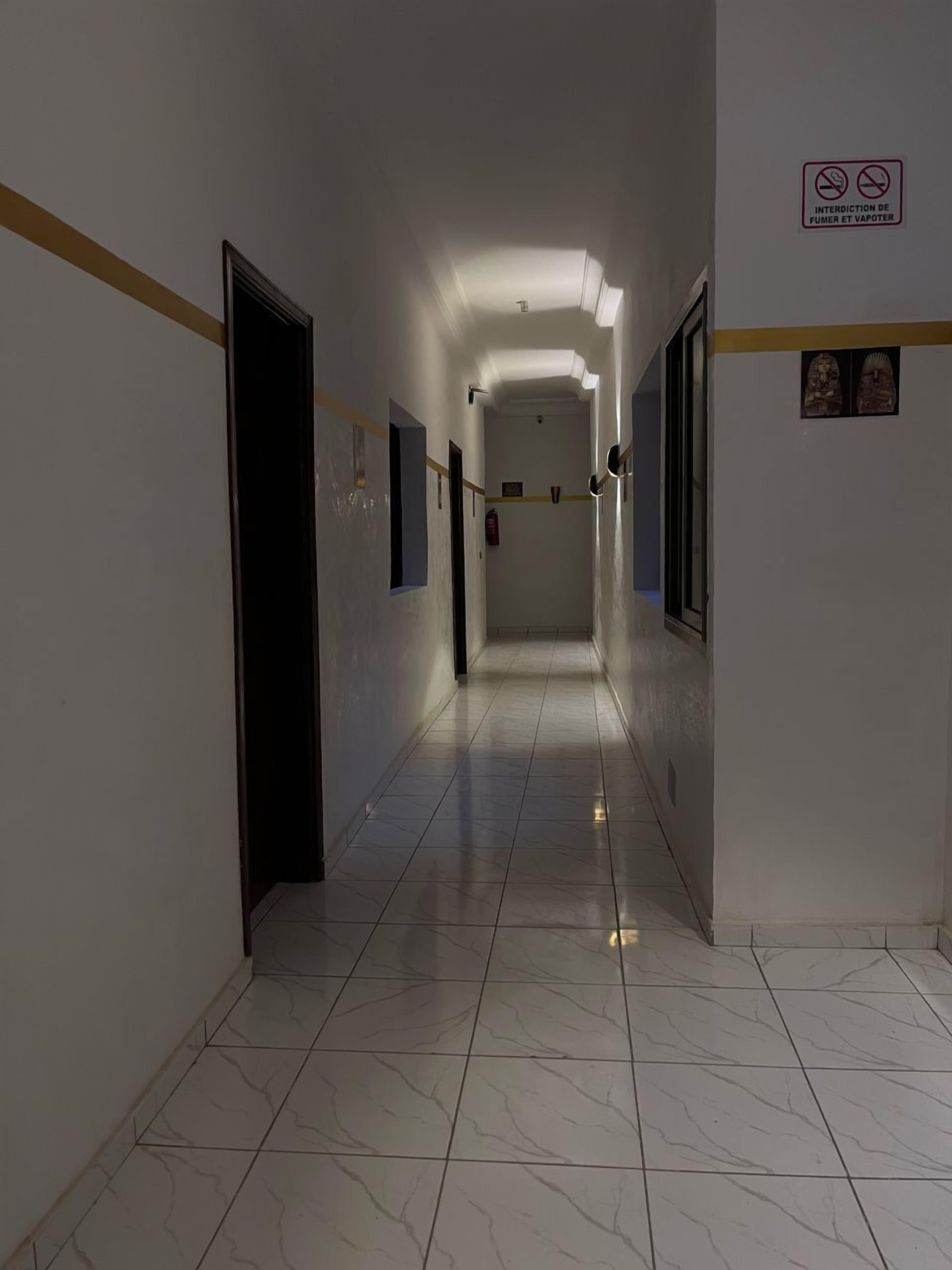 A long, narrow hallway with white walls and tiled floors. There are doors on both sides, and a sign prohibiting smoking and vaping is visible on the right wall.