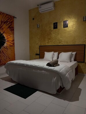 Ett hotellrum med en säng, vita sängkläder, en hoprullad handduk, en luftkonditionering och en vägg med en intressant yta.