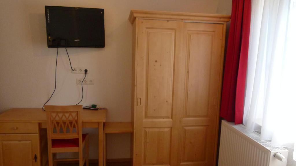 A room with a wooden desk, a chair, a television mounted on the wall, and a wardrobe.