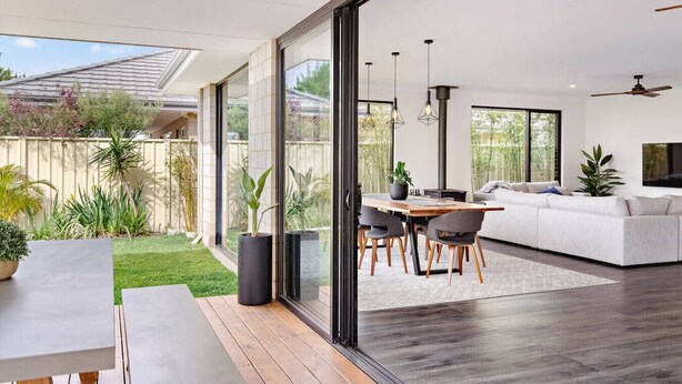 A modern living room with a large sliding glass door leading to a backyard. There is a dining area with a table and chairs, a sofa, and a ceiling fan. - Mainbreak Dunsborough – Stylish Coastal Retreat (Dunsborough)