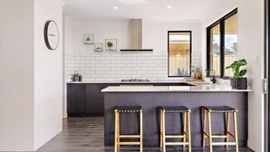 A modern kitchen with a white tiled backsplash, a central island with stools, a large window, and a clock on the wall. - Mainbreak Dunsborough – Stylish Coastal Retreat (Dunsborough)