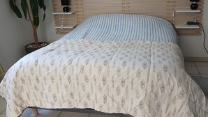 Ein Bett mit gestreiftem Kopfkissen, ein Nachttisch mit Lautsprecher, ein Wandregal und ein Topfpflanze.