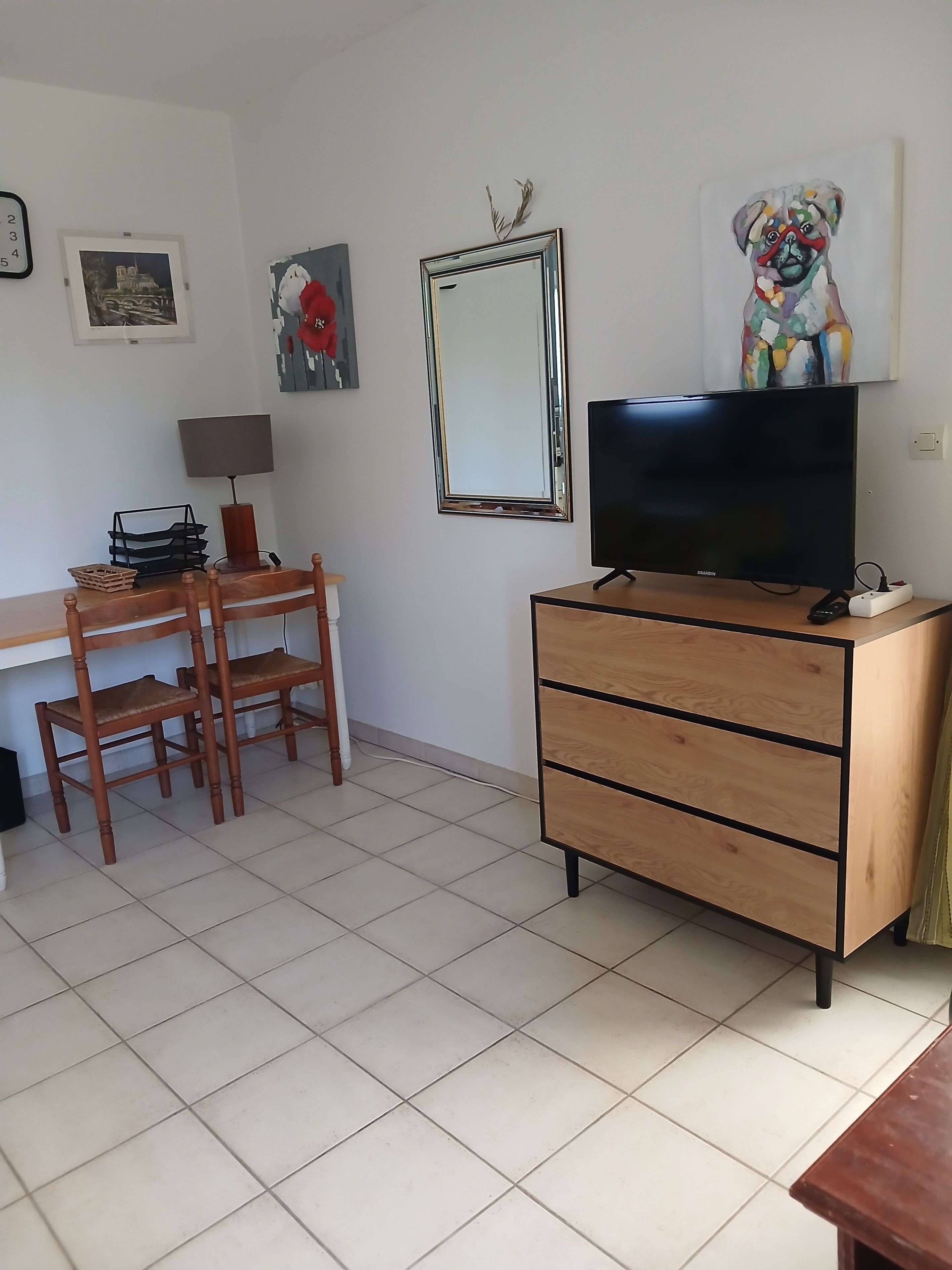 Ein Zimmer mit Schreibtisch, zwei Holzstühlen, einem Fernseher auf einem Holzschrank, einem Spiegel und Wandbildern.