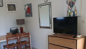 Ein Zimmer mit Schreibtisch, zwei Holzstühlen, einem Fernseher auf einem Holzschrank, einem Spiegel und Wandbildern.