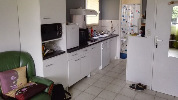 Eine kleine Küche mit weißen Schränken, einer Mikrowelle, einem Toaster und einem Spülbecken. Ein grüner Stuhl mit Blumenkissen und eine schwarze Tasche stehen auf dem Boden.