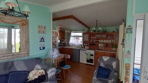A cozy living room with a sofa, a fan, and a small dog. A kitchen with wooden cabinets and a dining area is visible in the background. - 3 Bed Chalet - Sleeps 6 - Parking - Garden - Wifi (Great Yarmouth)