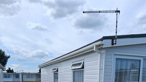 A white mobile home with a satellite dish, a small porch with a bench, and a potted plant. - 3 Bed Chalet - Sleeps 6 - Parking - Garden - Wifi (Great Yarmouth)