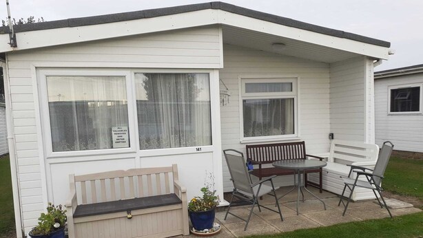A small, single-story mobile home with a porch, a bench, a table, and two chairs. - 3 Bed Chalet - Sleeps 6 - Parking - Garden - Wifi (Great Yarmouth)