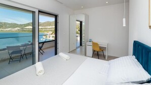 A modern hotel room with a large bed, a desk, and a balcony with a view of the sea and hills.