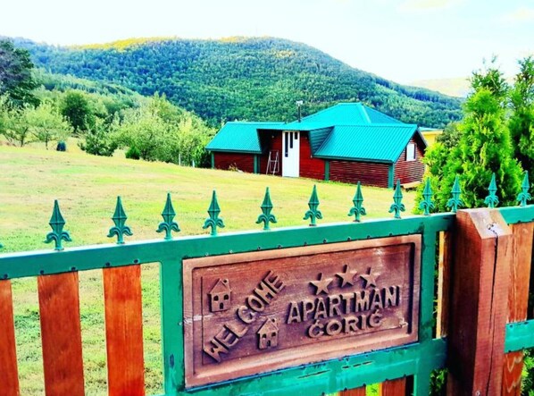 A wooden sign with "WELCOME APARTMANS CORIG" and a house in the background.