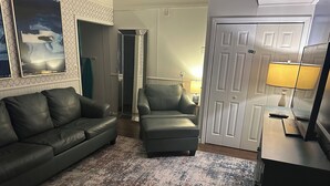 Una sala de estar con un sofá de cuero gris, un sillón gris, un tapete estampado, una puerta blanca y un televisor sobre un mueble.