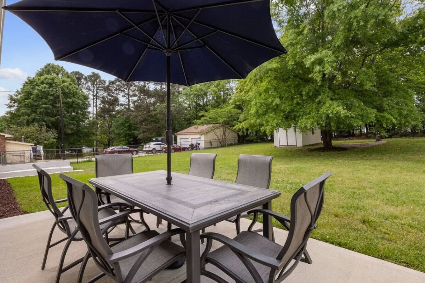 An outdoor patio with a table and chairs, a large umbrella, and a grassy area with trees and a fence in the background.