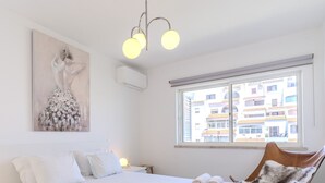 Ein Schlafzimmer mit Bett, einem Gemälde, einem Fenster mit Blick auf Gebäude und einer Deckenleuchte.