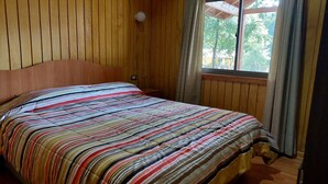 Ett rum med en väggpanel av trä, en säng med ett randigt överkast, ett fönster med gardiner och en takkonstruktion av trä.