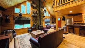 A cozy cabin interior with a wooden ceiling, stone walls, a fireplace, a leather sofa, a dining area, and a kitchen.
