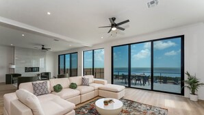 A spacious living room with a large L-shaped sofa, a round coffee table, and a view of the ocean through large windows. - 4,000 SF Waterfront Home w/ Pool, Sleeps 22 (Port Isabel)