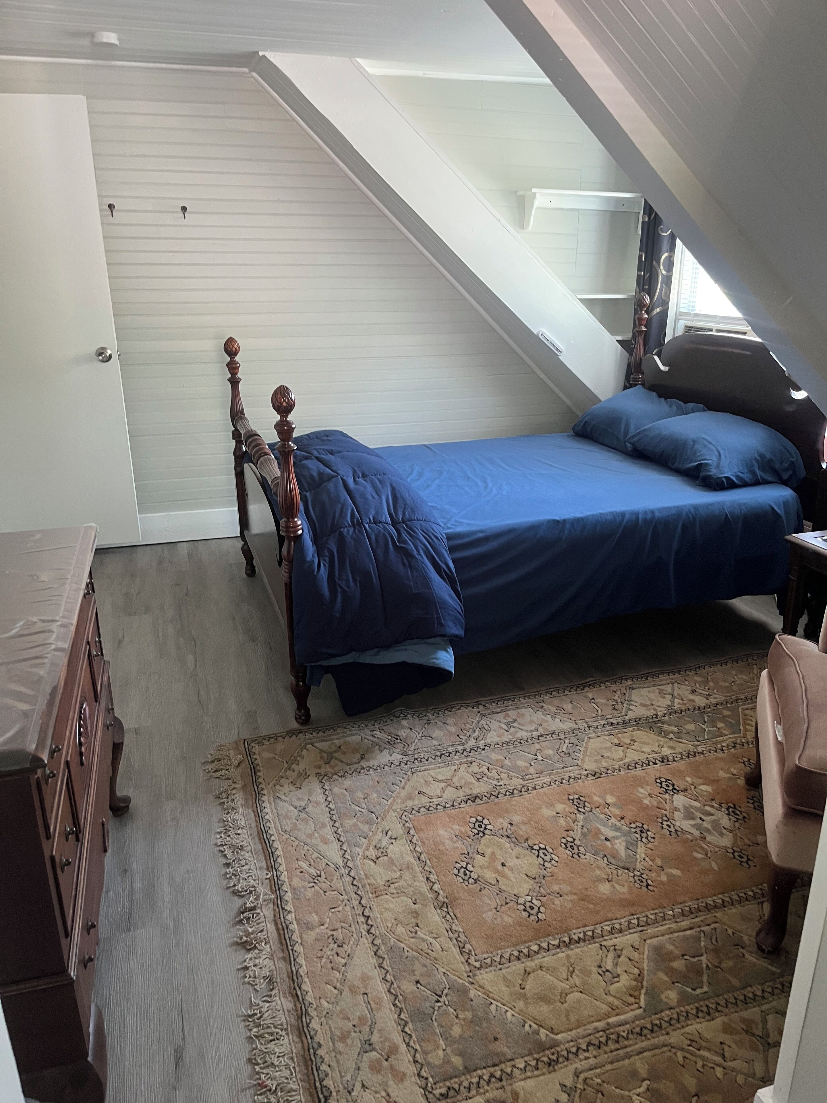 A bedroom with a sloped ceiling, a bed with blue bedding, a wooden dresser, and a patterned rug.
