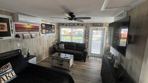 Ein Wohnzimmer mit einer Couch, einem Couchtisch und einem Fernseher. Eine Tür mit der Aufschrift 'LAKE' und ein Deckenventilator sind vorhanden.